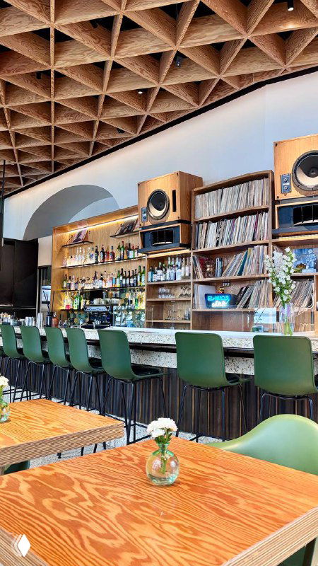Барная зона Flaner с открытыми полками, виниловыми пластинками и аудиотехникой, длинная стойка и ряд зелёных барных стульев.