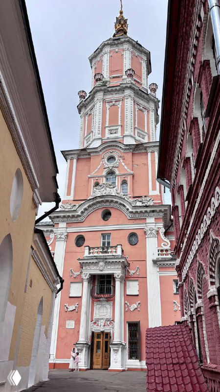 Розово‑белый фасад Храма архангела Гавриила XVIII века, снятый в перспективе между домами; видно лепнину, вход и верхний купол с крестом.