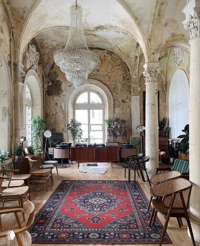 Большой зал с высокими арками, хрустальной люстрой и старинным ковром на полу, ощущение запущенной, но красивой архитектуры и света.