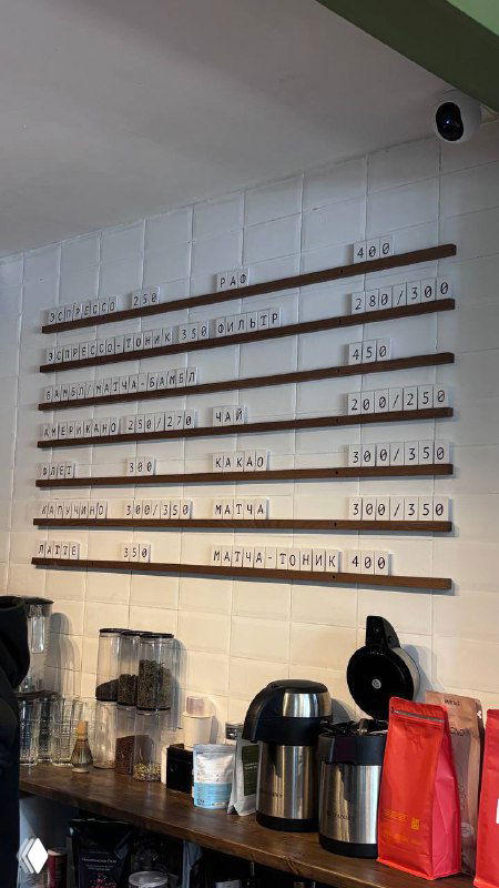 Стена меню и стойка в кофейне: деревянные рейки с названиями, чайники и емкости — атмосфера небольшого локального заведения.