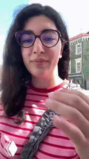 Селфи автора у окна кафе: она демонстрирует жест, на заднем плане видны фасады соседних зданий и дневной свет.