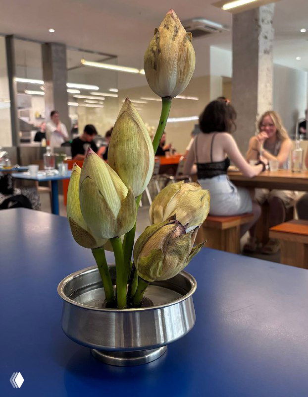 Небольшая композиция с букетом бутонов на столе кафе; видно пространство зала и посетителей на заднем плане в интерьере Zoe.