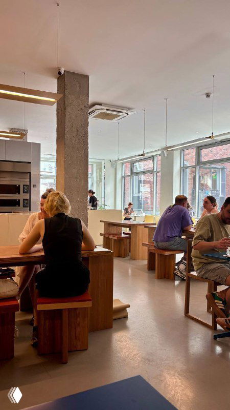 Интерьер пекарни Zoe: деревянные столы и стулья, большие окна и дневной свет, посетители за столиками — чистый современный стиль.