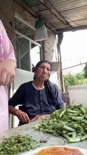 Пожилая женщина сидит за столом на веранде сельского дома, рядом стопка свежесорванной стручковой фасоли и посуда, передаётся деревенская атмосфера.