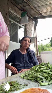 Бабушка за столом во дворе села Схвилиси, рядом стопка свежих стручков фасоли, видно крыльцо и зелёные окрестности.