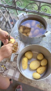Подготовка картофеля: чистка и промывка клубней в большой кастрюле, видна рабочая поверхность и руки у плиты на свежем воздухе