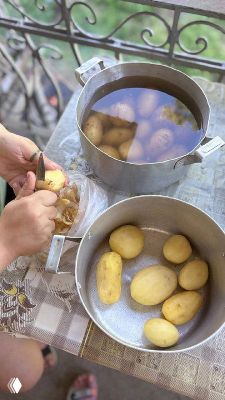 Большая кастрюля с очищённой картошкой на столе на улице, руки рядом чистят картофель, вид деревенской кухни и подготовки к запеканию в фольге.