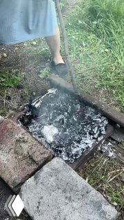 Разгоревшиеся угли в самодельном мангале, вокруг пепел и дым, видно рабочее место на земле в саду — атмосфера деревенского очага.