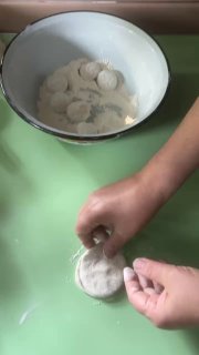 Рука раскатывает кружок теста и формирует приплюснутую заготовку: видна техника формирования оболочки хинкали в домашней кухне.