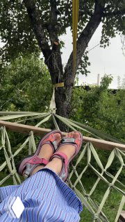Ноги в сандалиях в гамаке под деревом: расслабленная поза, вид на сад и полотно гамака, тёплая летняя атмосфера отдыха на даче.