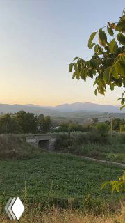 Панорамный вид на сельскую долину с полями, мостом и дальними холмами на закате: тихая летняя сельская сцена с мягким светом.