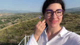 Портрет улыбающейся женщины в очках на фоне холмов и долины; в руке небольшой полевой цветок, тёплое солнце позади.