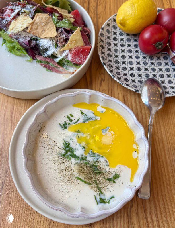 Глубокая миска супа танов абур с белой и жёлтой эмульсией, зеленью и перцем; рядом тарелка с лимоном и сладкими помидорами на столе.