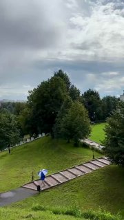 Пейзаж парка усадьбы Дубровицы: дорожка по склону, посетитель с зонтом, лужи после дождя и зелёные массивы деревьев.