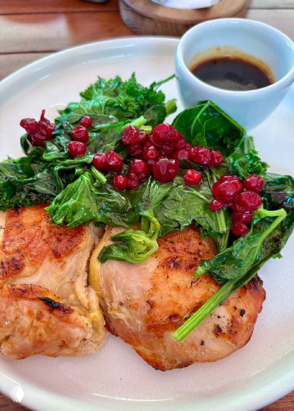 Крупный план тарелки с сочной запечённой курочкой, зеленью, яркими красными ягодами и картофельным гарниром на деревянном столе в ресторане.