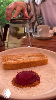 Подача десерта и рук официанта с чайником на заднем плане — кадр отражает атмосферу сервиса и тёплую обстановку на веранде.