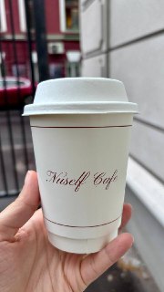 Бумажный стакан с логотипом Nuself Cafe в руке на улице; крупный план стакана с крышкой и фирменной надписью кафе.