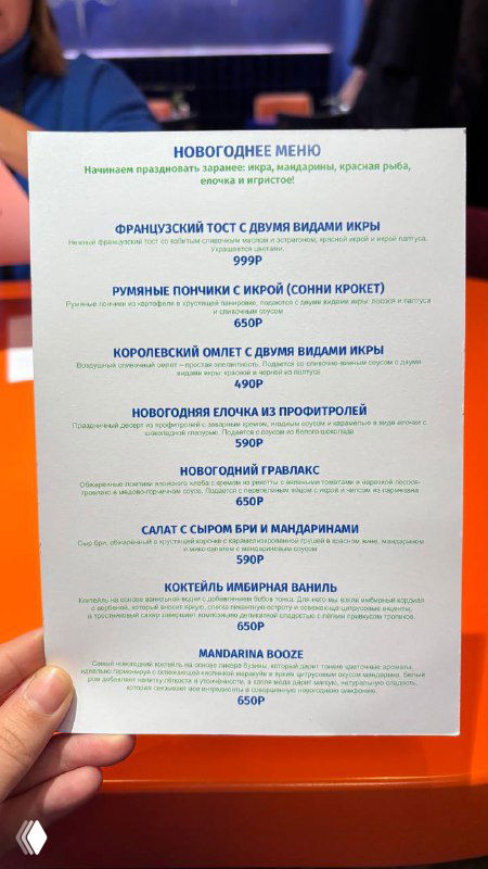 Рука держит распечатанное новогоднее меню кафе Скворец с перечнем праздничных блюд и цен — листовка сезона на столе