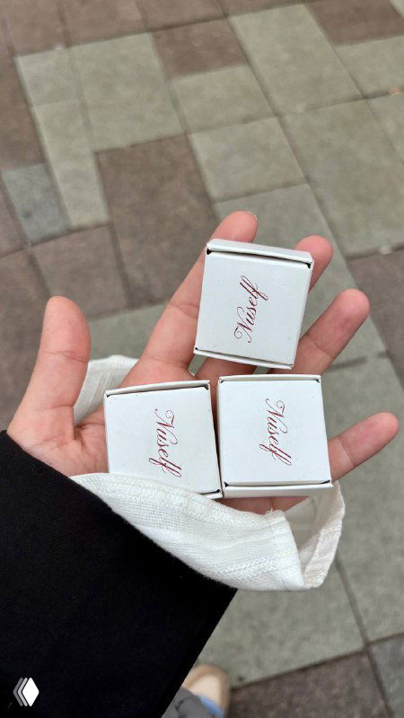 Рука держит маленькие коробочки конфет с логотипом Nuself — три аккуратные коробочки птичьего молока, видна текстура упаковки и рука на фоне улицы.