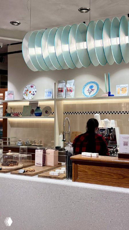 Кофейный прилавок и витрина с тарелками и декором: бариста за стойкой, стильный интерьер с пастельными акцентами и открытой зоной приготовления напитков.