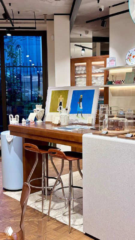Барная стойка и рабочая зона кафе Nuself: высокие стулья, столешница с аксессуарами, произведения искусства и окно на улицу в глубине интерьера.