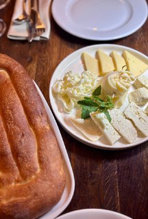Тарелка с несколькими видами белых сыров, маслом и горячей лепёшкой на деревянном столе в уютном интерьере ресторана, крупный план.