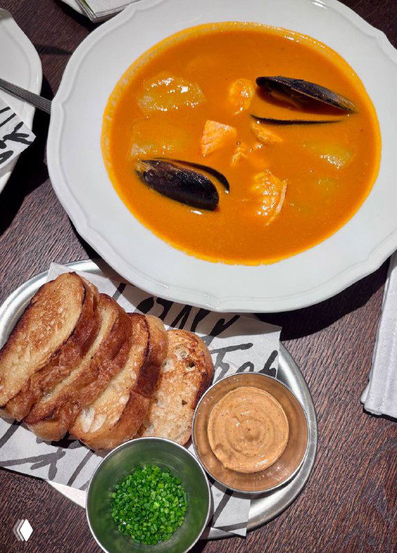 Крупный план буйабеса в белой тарелке с мидиями, гренками и соусом — насыщенная апельсиновая похлёбка в ресторанной подаче.