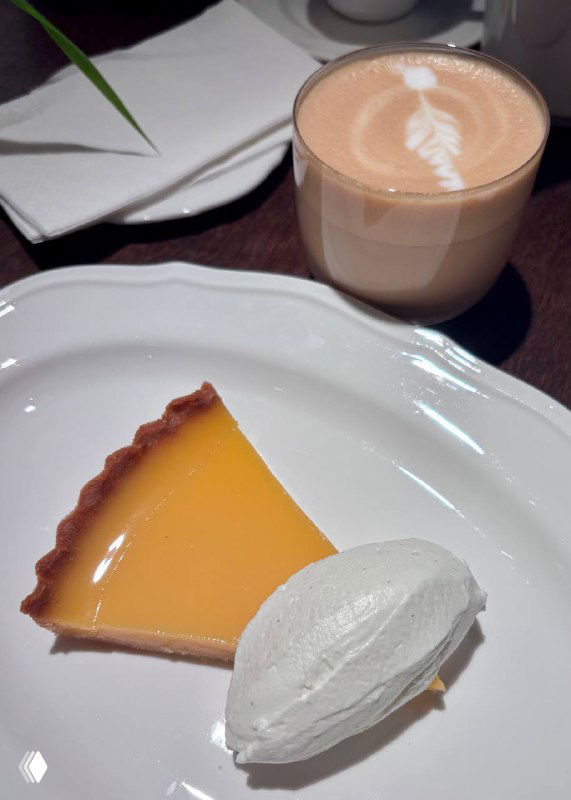 Лимонный тарт на белой тарелке с шаром взбитого крема и чашкой кофе рядом — десерт с выраженной кислинкой и нежной текстурой.