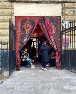 Вход в небольшой турецкий спот: широкий проём, декоративные ковры как занавеска и несколько сидящих людей у порога.