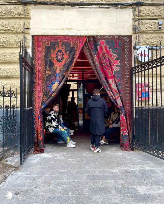 Вход в Mini Turkish Spot на Тверской с подвешенными красочными турецкими коврами; у прохода сидят гости, виден городской поток.