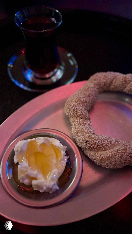 Чай и симит на подносе: бублик в плотной кунжутной корочке, рядом мисочка с густым каймаком и медом, тёплая вечерняя подача.