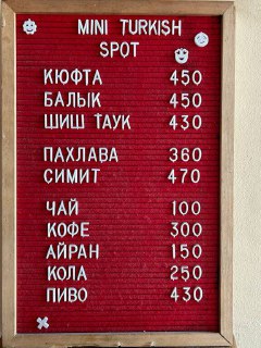 Вертикальная табличка-меню с красным фоном и белыми буквами: перечень блюд и цен для посетителей на входе в спот.