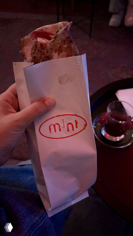 Завёрнутая шаурма/балык в фирменном пакете Mini, на переднем плане рука держит еду, рядом стакан турецкого чая в традиционной форме.