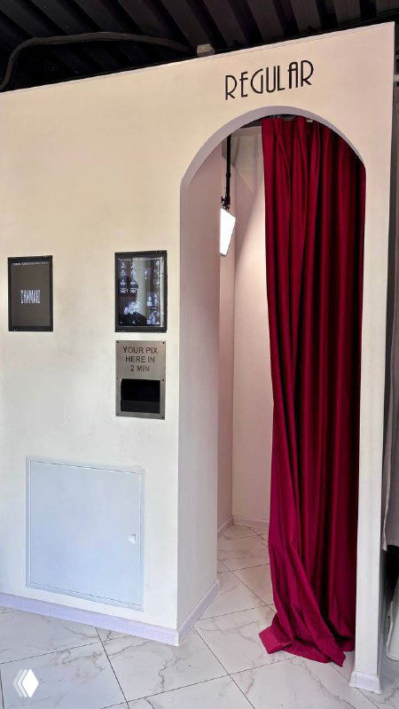 Вход в кабину фотобудки: белая стена, автомат для печати и открытая красная занавеска — интерьер студии Photopix в центре города.