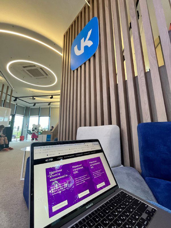 Интерьер коворкинга VK.DOM: ноутбук на столе, фоновая стеновая панель с логотипом VK, мягкие кресла и современное круглое потолочное освещение.