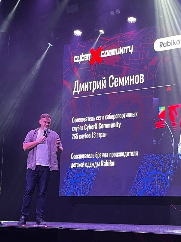 Докладчик на сцене у большого экрана с надписью «Дмитрий Семинов»: мужчина говорит в микрофон на фоне брендированных слайдов и сценического освещения.