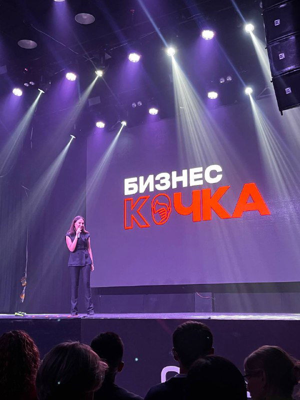 Женщина на сцене рядом с ярким экраном и логотипом «БИЗНЕС КОЧКА»: прожекторы, сцена и силуэты зрителей в переднем плане.