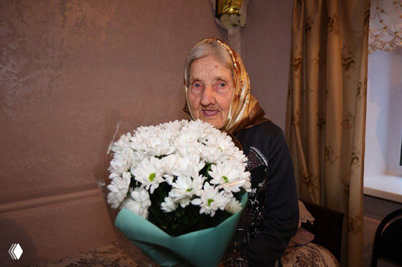 Пожилая женщина — Прасковья Николаевна Болдина — с большим букетом белых хризантем в домашнем интерьере, портрет к столетию.