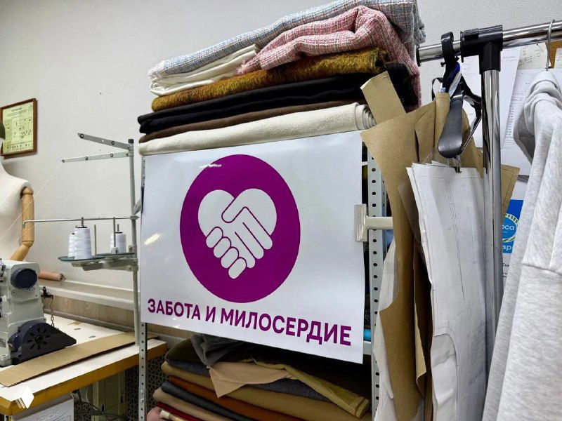 Вывеска «Забота и милосердие» в швейной мастерской на фоне рулонов ткани и оборудования для пошива, рабочая зона видна.