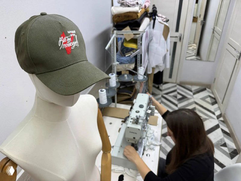 Манекен с кепкой и швея за промышленной швейной машиной в мастерской проекта адаптивной одежды, вид рабочего процесса.