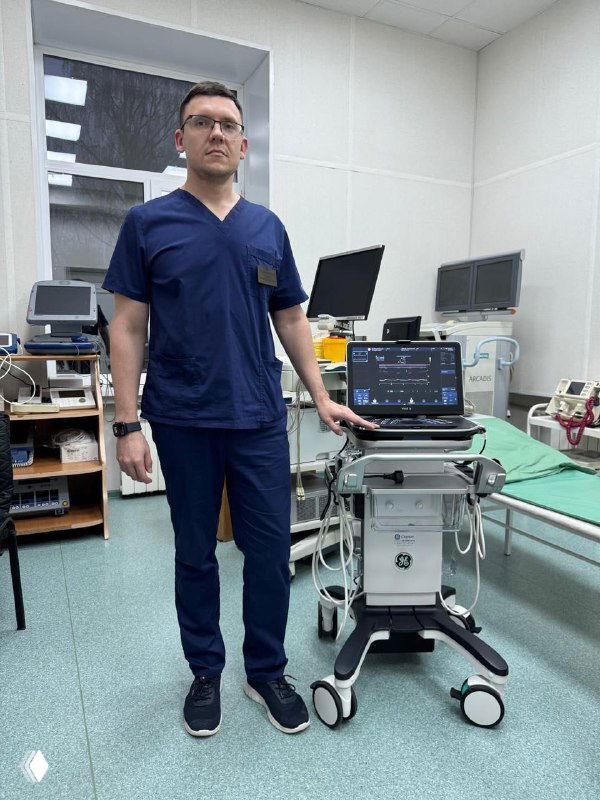 Врач в синей форме рядом с новым высококлассным внутрисердечным УЗИ-аппаратом на мобильной тележке в кардиодиспансере.