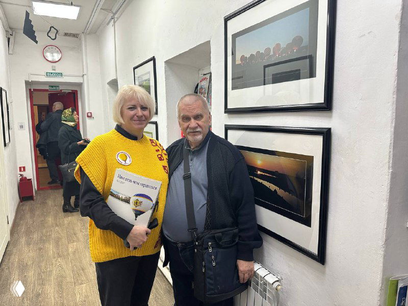 Два человека на открытии выставки в Рязанском Фотодоме рядом с оформленными фотоработами из серии «Небо Рязани», видна галерея и рамы на стене.