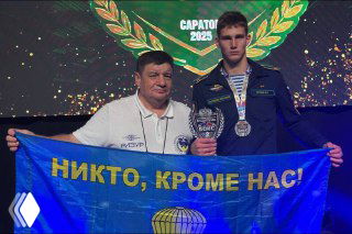 Михаил Астахов в спортивной форме с медалью и кубком на фоне сцены соревнований, держит синий флаг с надписью «Никто, кроме нас!», знак победы на турнире.