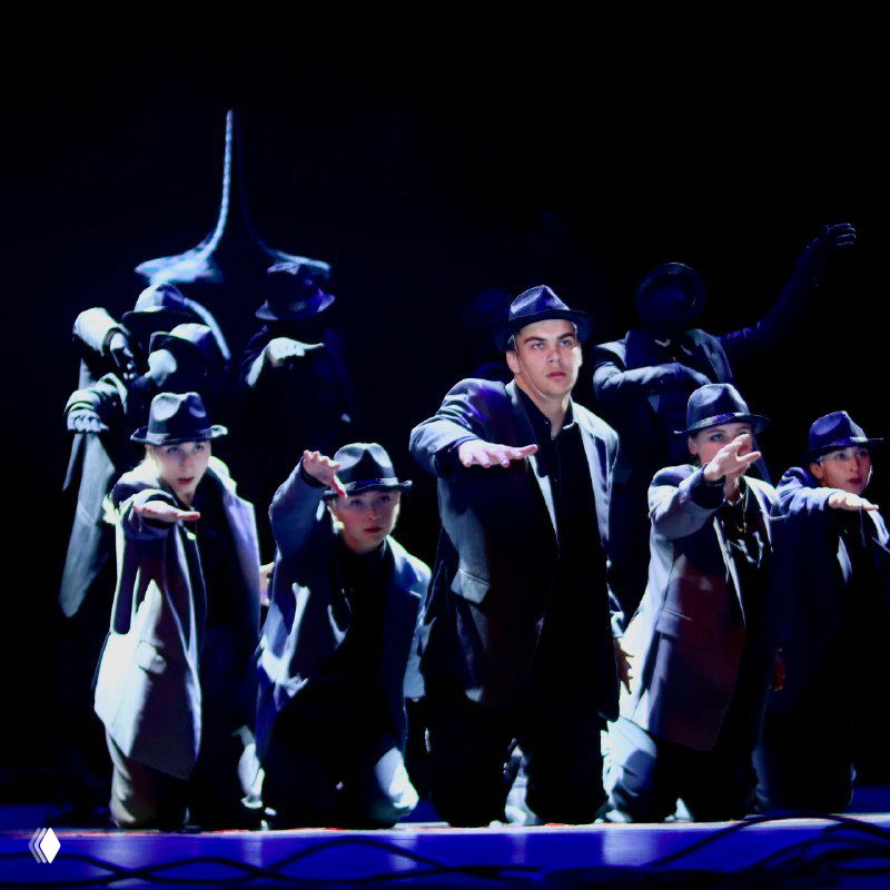 Хореографический номер с мужской группой танцоров в стилизованных костюмах, драматическое освещение и выразительные позы участников на сцене.