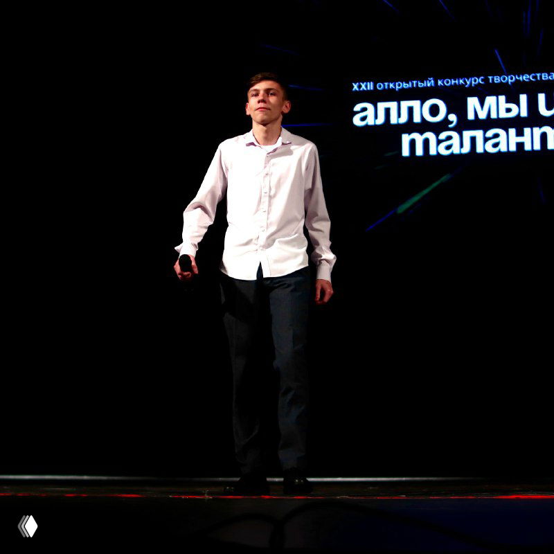 Солист в белой рубашке выступает с микрофоном на сцене фестиваля «Алло, мы ищем таланты!», видно проекцию с названием конкурса на заднике.