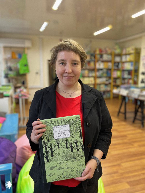 Женщина среднего возраста держит книгу «Про кабанов, бобров и выхухолей» на фоне книжных полок в интерьере магазина.