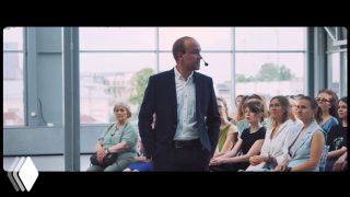 Мужчина в костюме выступает перед собравшейся аудиторией в зале; сцена похожа на презентацию или лекцию, люди слушают в зале.