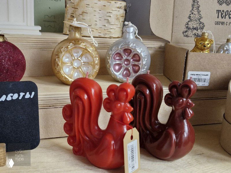 Крупный план: яркие фигурные ёлочные игрушки в виде красных петухов и других сувениров на полке в магазине «Резиденции традиций», бирки цен видны на изделиях.