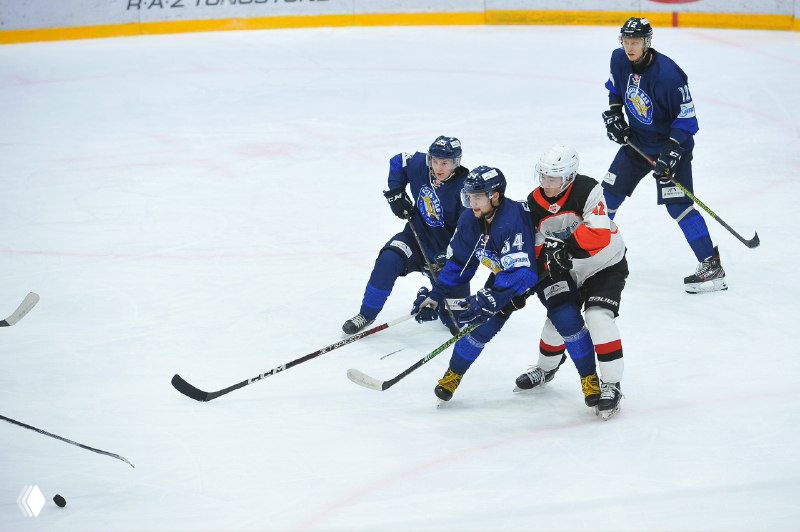 Хоккеисты в синих свитерах команды «Рязань-ВДВ» активно борются за шайбу на льду дворца спорта «Олимпийский», видны борта и часть трибун с болельщиками