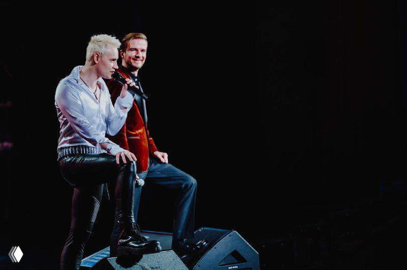 Два артиста на сцене Кремлёвского дворца: сидят у края сцены с микрофонами, видна часть оборудования и тёмный задник.